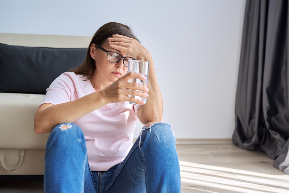 Sad middle aged woman sitting at home on the floor with glass of water.