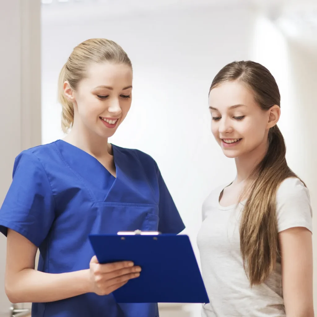 A smiling healthcare professional in blue scrubs reviews a clipboard with a young female patient.