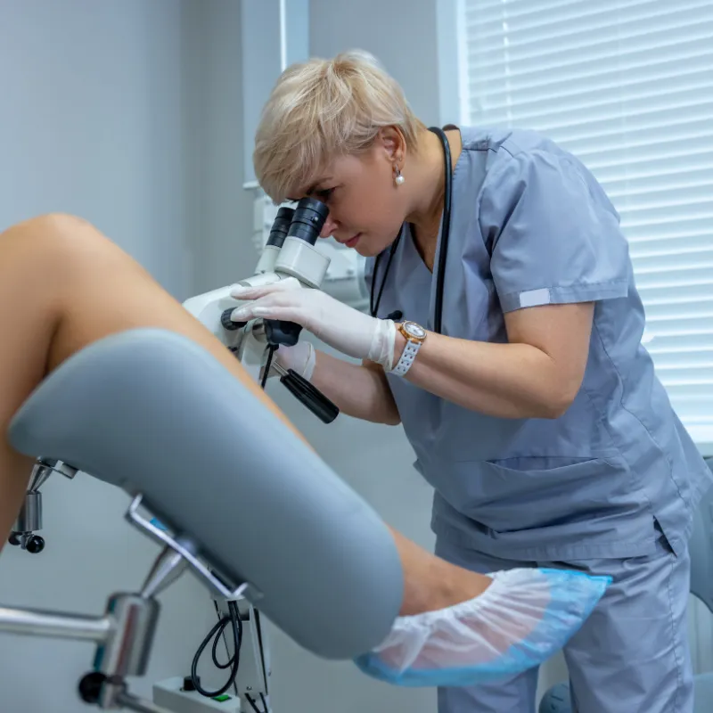 A gynecologist in scrubs uses a colposcope during a patient examination.