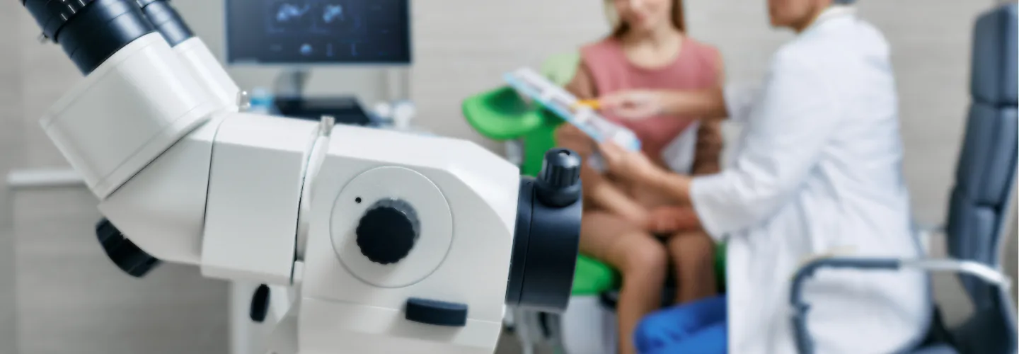 A colposcope in focus with a doctor and patient consulting in the blurred background.