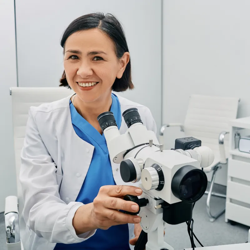 A smiling female gynecologist in a white lab coat sits next to a colposcope in a medical office.