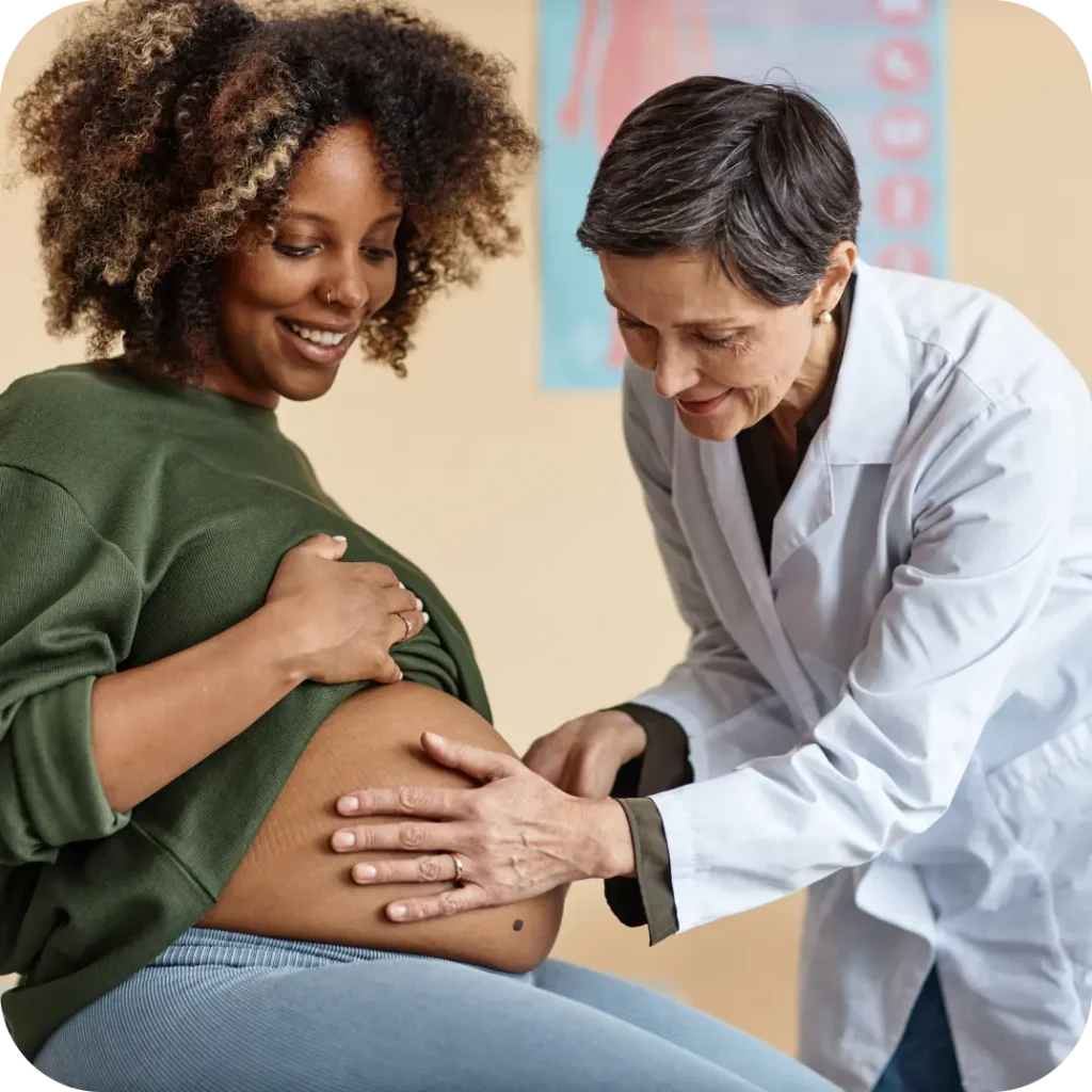 A smiling obstetrician gently examines a pregnant Black woman's belly during a prenatal checkup.