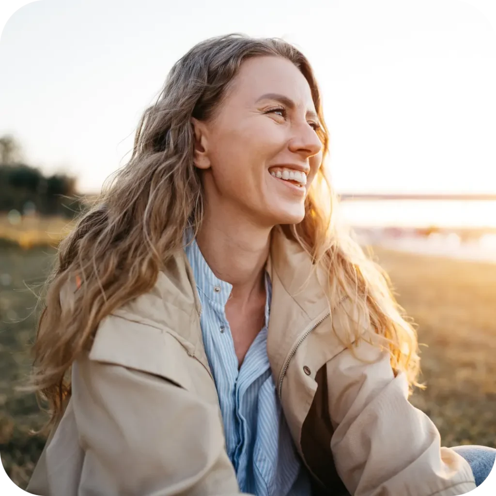 A happy, smiling woman with long wavy hair sits outdoors during golden hour.