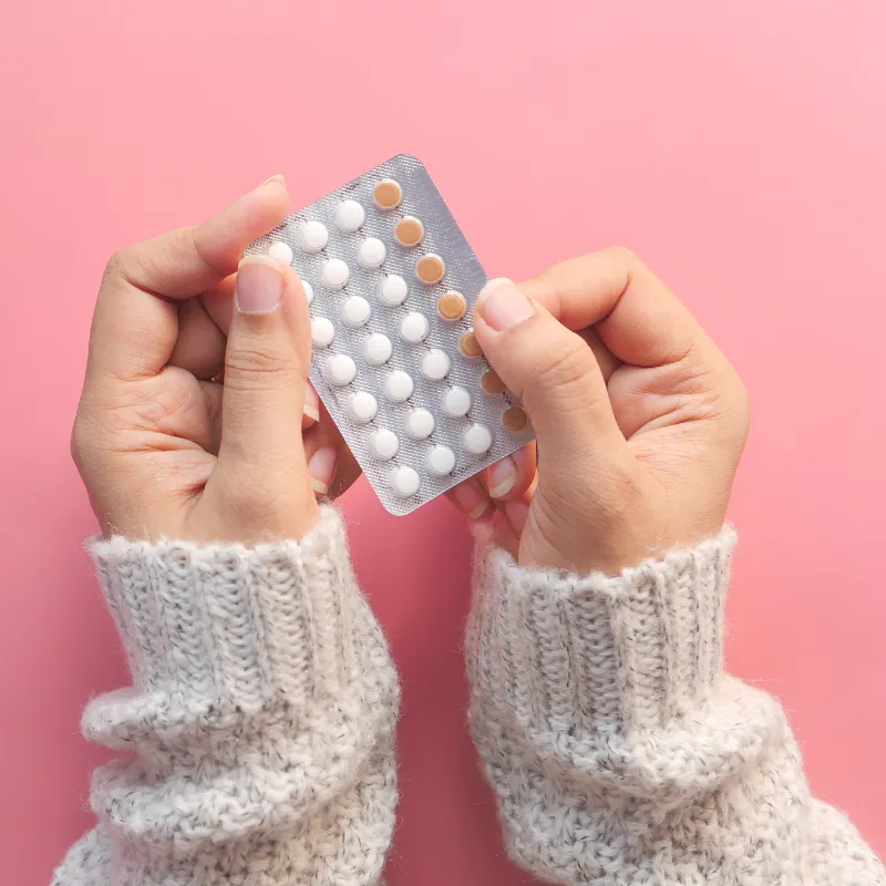 Hands holding a blister pack of birth control pills against a pink background.