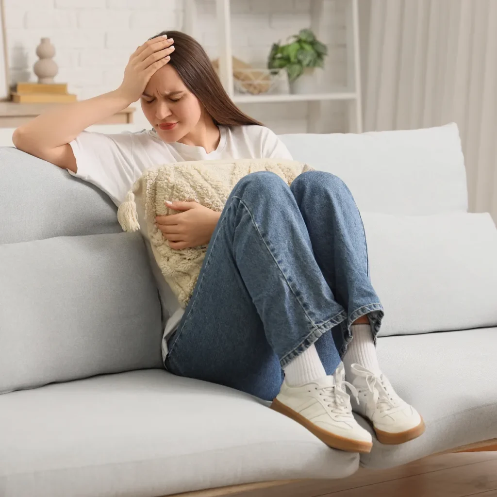 A woman sits on a couch grimacing in pain, holding her head and clutching a pillow to her stomach.