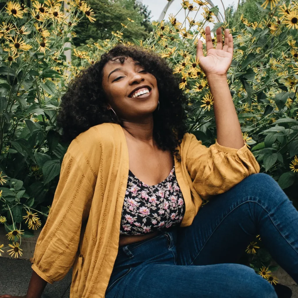 A happy Black woman with curly hair smiles and waves, sitting in front of vibrant yellow flowers.
