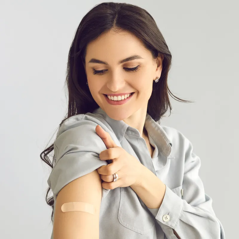 A smiling woman looks down at an adhesive bandage on her upper arm after receiving a vaccine.