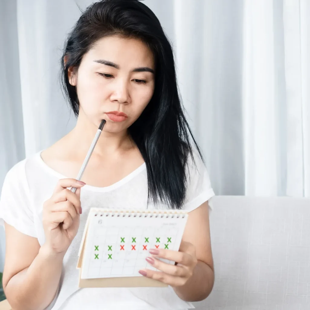 A woman looks thoughtfully at a calendar marked with red and green Xs, tracking her menstrual cycle.