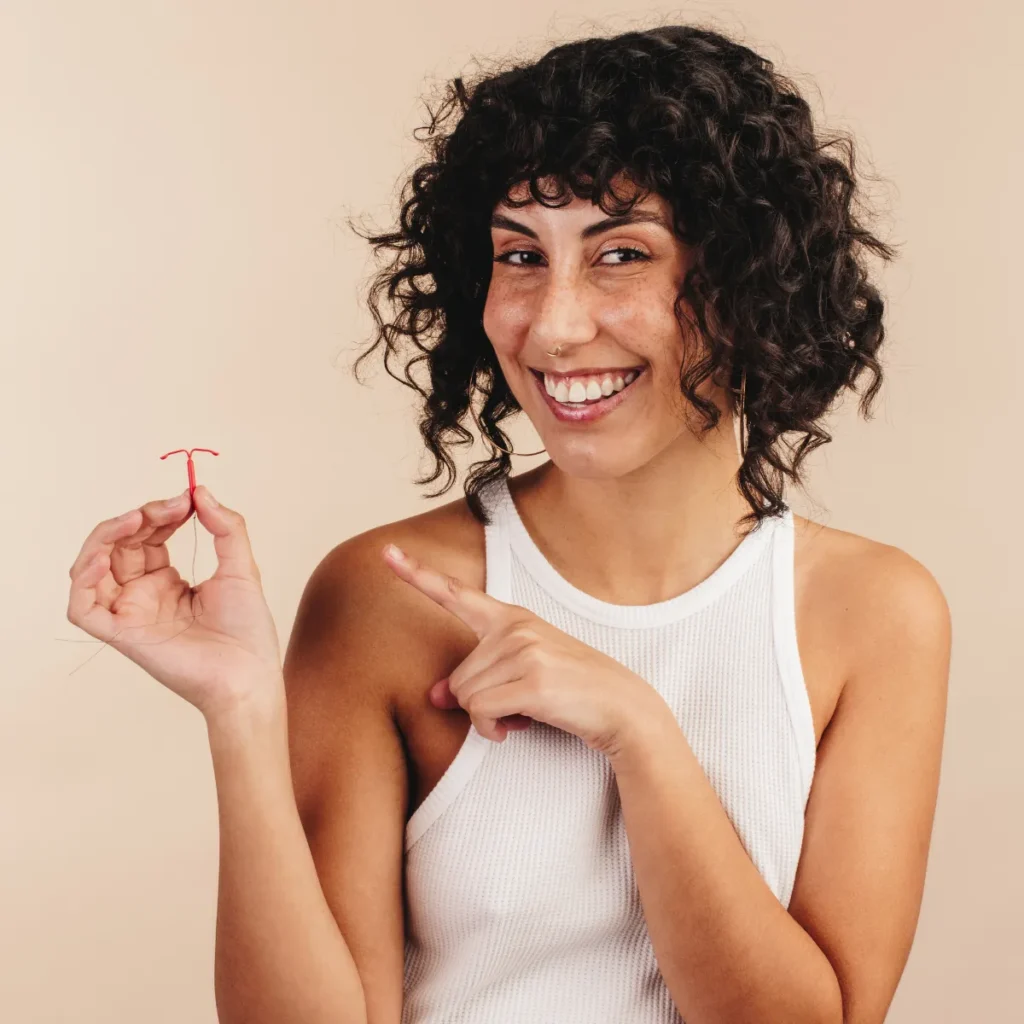 A smiling woman points to a red intrauterine device (IUD) held in her hand.