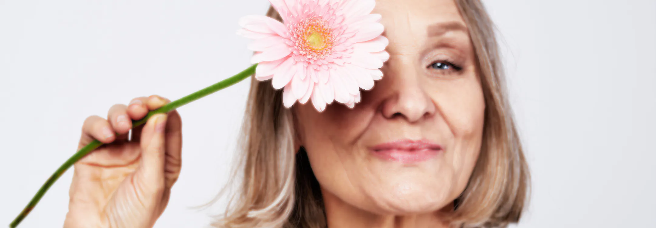 A smiling older woman holds a pink daisy over her eye.