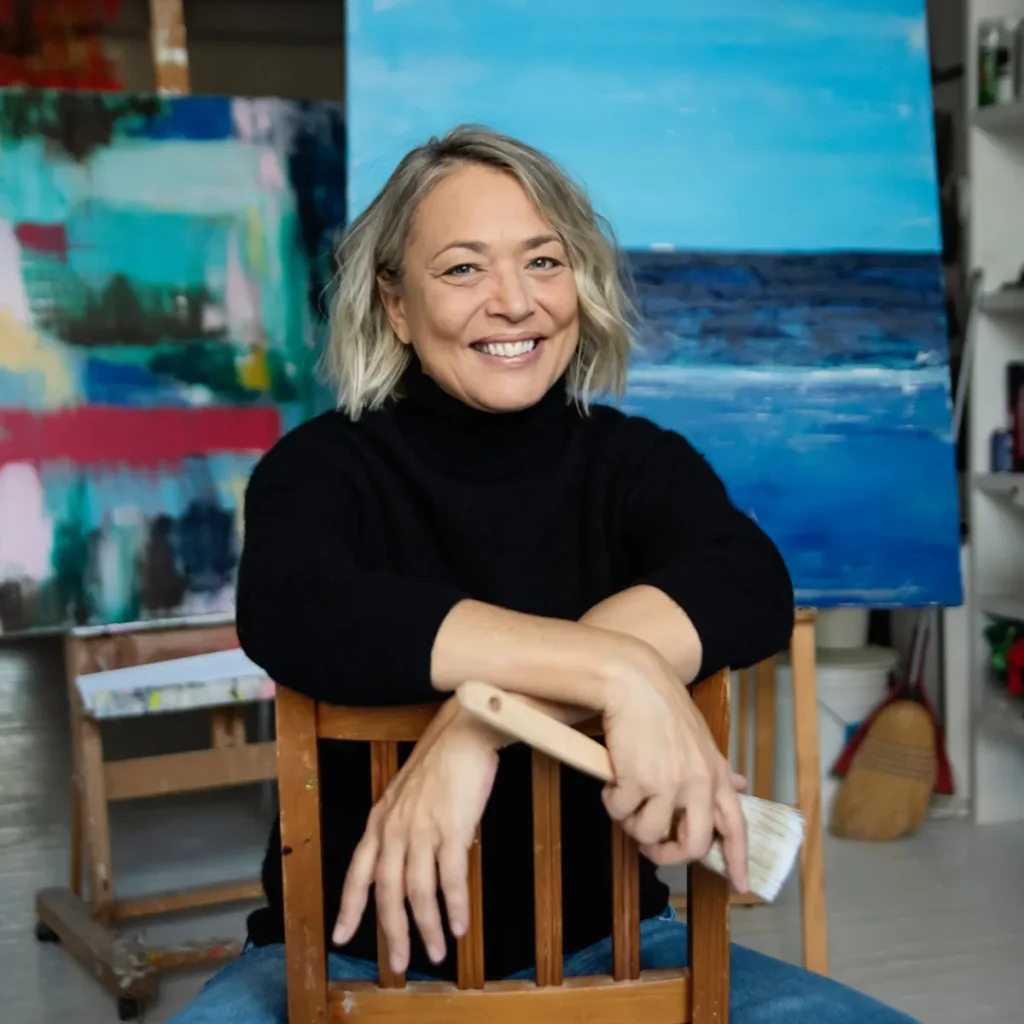 A smiling woman sits in an art studio holding a paintbrush, with colorful paintings in the background.