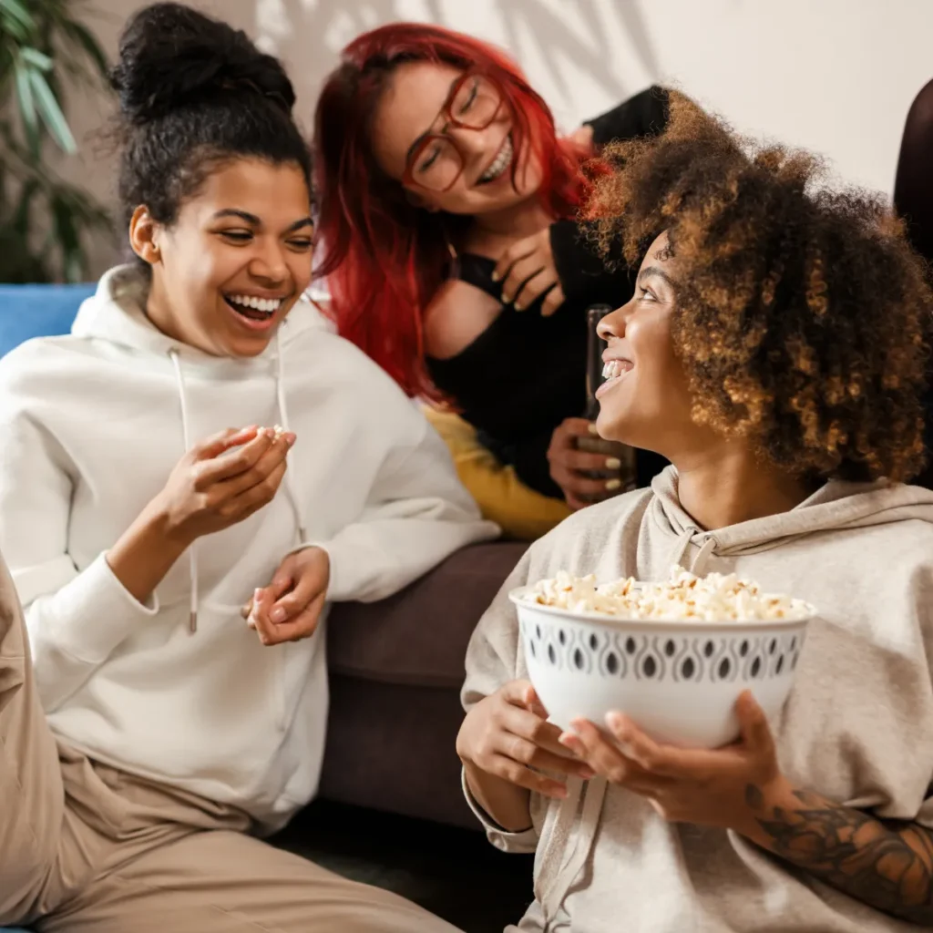 Three diverse young women smiling and laughing together while sharing a bowl of popcorn.
