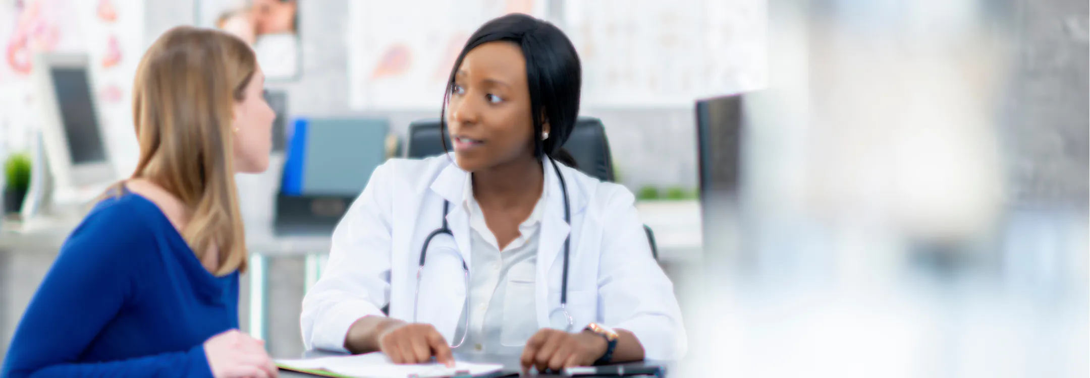 A female doctor in a white lab coat and stethoscope speaks with a patient during a medical consultation.
