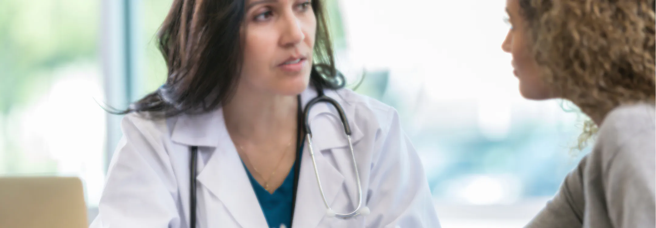 A female doctor in a white coat speaks with a female patient during a consultation.