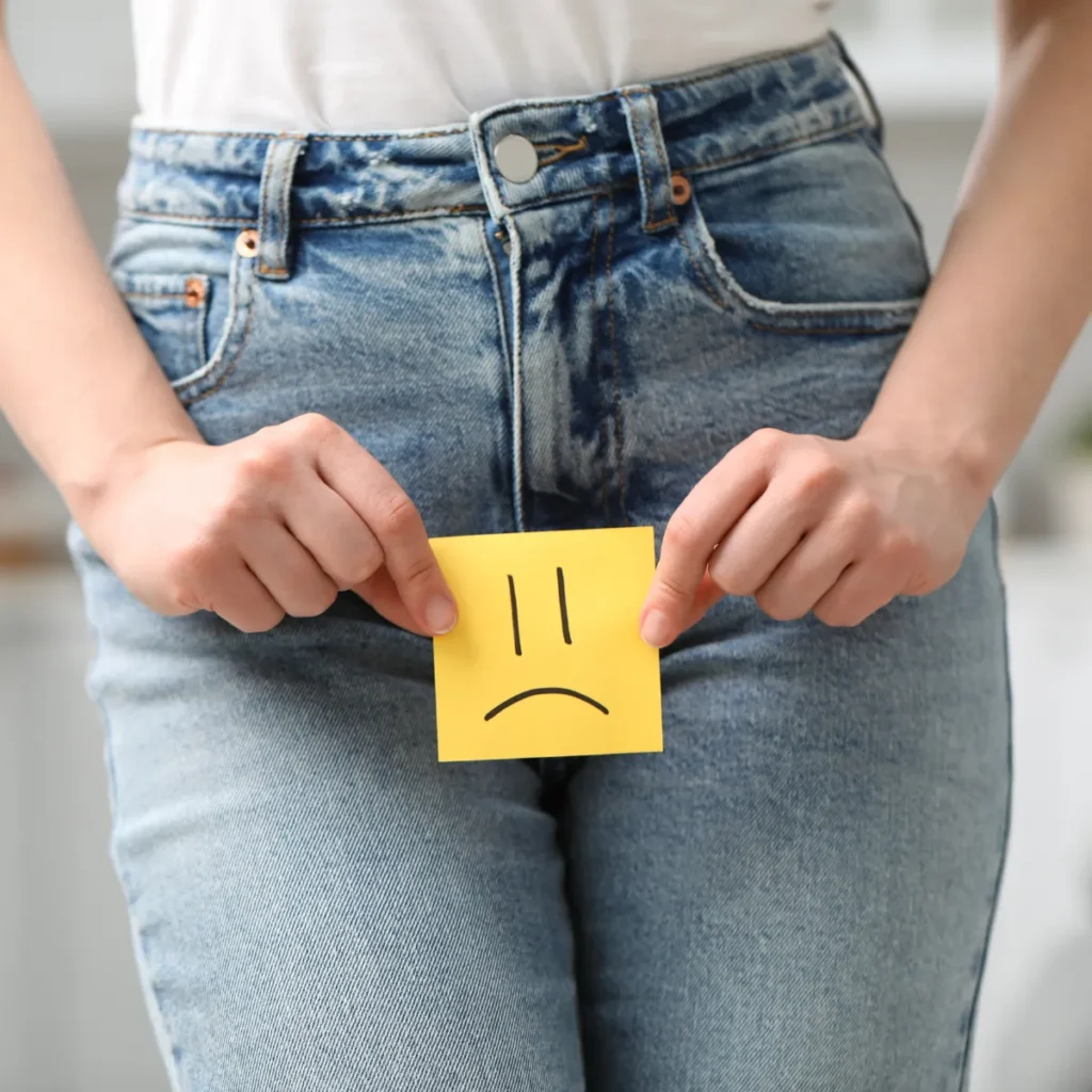 A woman holding a yellow sticky note with a sad face over her pelvic area, representing pain or discomfort.