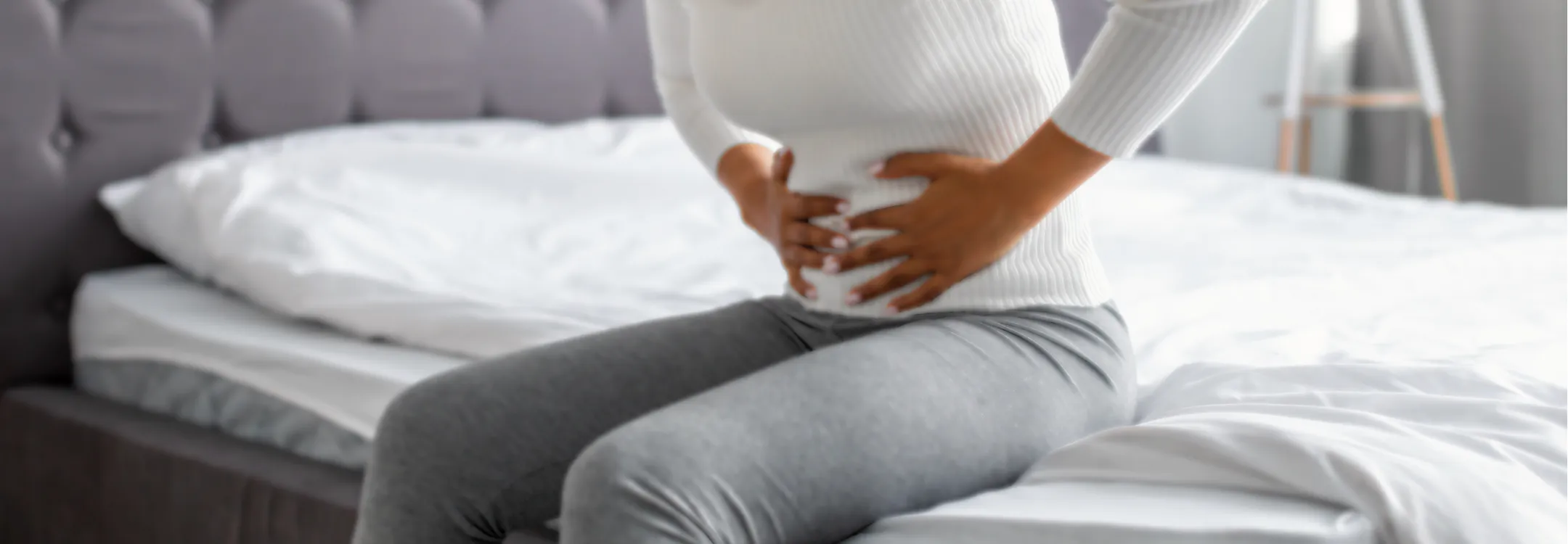 A woman sitting on a bed clutches her abdomen, experiencing pelvic or abdominal pain.