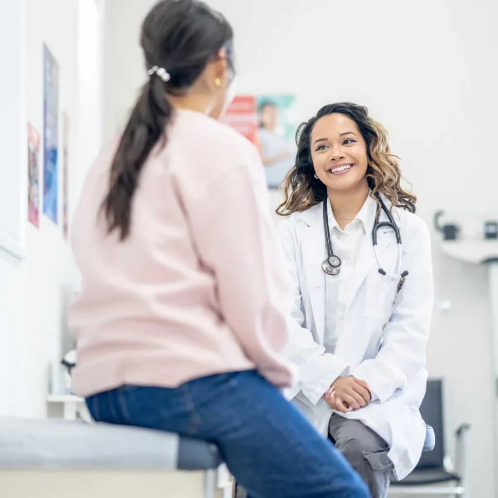 A smiling female doctor consults with a patient sitting on an exam table.