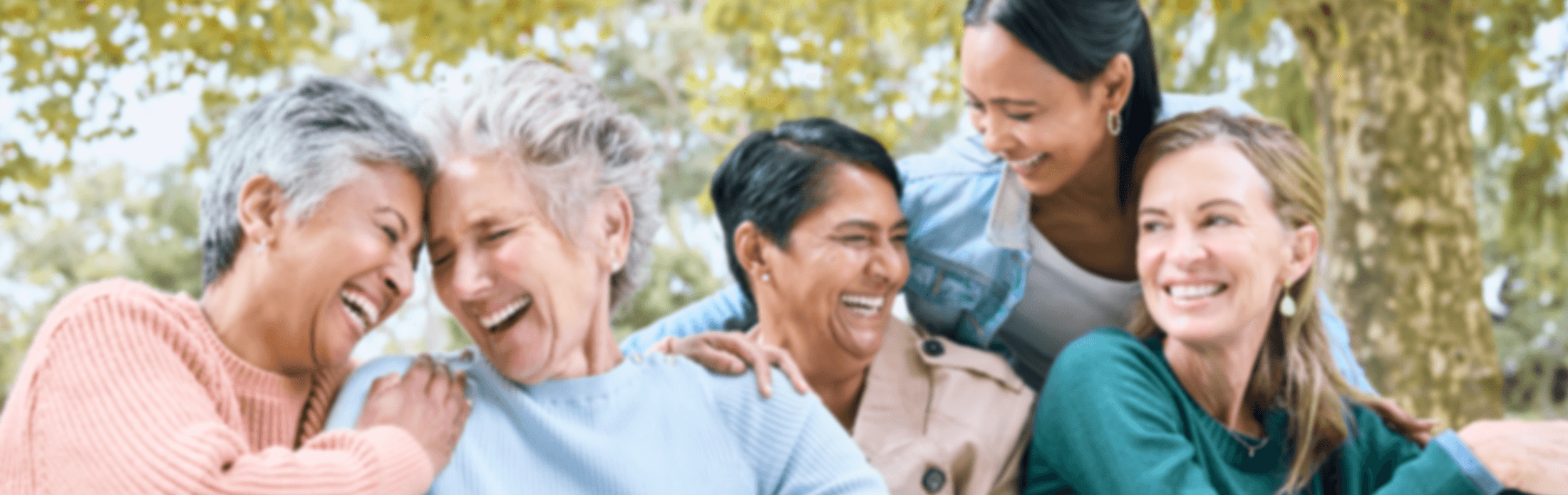 Five diverse, multi-generational women smiling and laughing together outdoors.