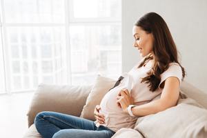 Smiling pregnant young woman holding headphones at her belly while sitting on a couch